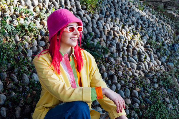 Urban spring street fashion look. Vanilla Girl. Kawaii vibes. Candy colors design. Bucket hat trend. Young woman with pink hair and sunglasses in multicolor outfit on the stones and plants background. - Fotoğraf, Görsel