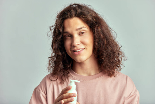 A young handsome smiling guy with long brown hair holds a bottle of a moisturizing beauty product in his hand. Caucasian man in the studio with healthy skin. Wellness, cosmetics and skin care for men - Foto, Imagen
