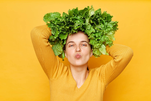 Green food lifestyle. Slimming weight loss. Care for wellness. Romantic woman with lettuce hair style with green vegetables on her head sending air kiss with pout lips isolated over yellow background - Фото, изображение