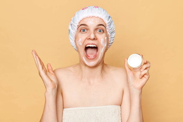 Body, skin care, cosmetics, beauty routine, treatment, bathroom, spa. Amazed woman wrapping in towel with shower cap on head cream in her hands isolated over beige background. - Photo, image