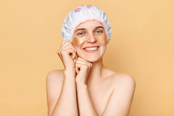 Healthy skin care routine. Cosmetic beauty secrets. Beauty product recommendations. Eye care essentials. Happy woman wearing shower cap with patches isolated over beige background - Zdjęcie, obraz