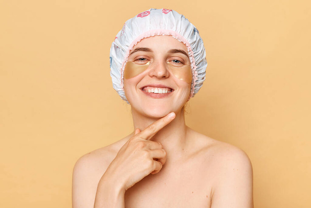 Perfect beauty regimen. Cosmetology expertise. Beauty patch application. Charming joyful woman wearing shower cap with eye patches isolated over beige background - Fotografie, Obrázek