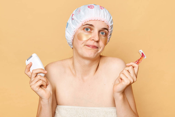 Confused puzzled woman wearing shower cap with eye patches isolated over beige background holding epilator and razor having doubtful expression choosing. - Photo, image