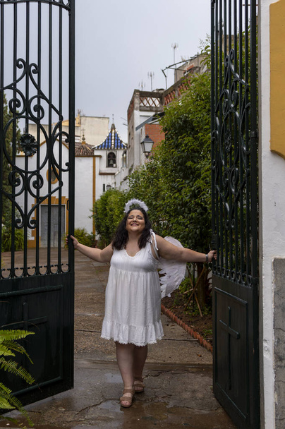 Caucasian woman with curly brown hair dressed as an angel with white dress and wings coming out of the hermitage - Photo, Image
