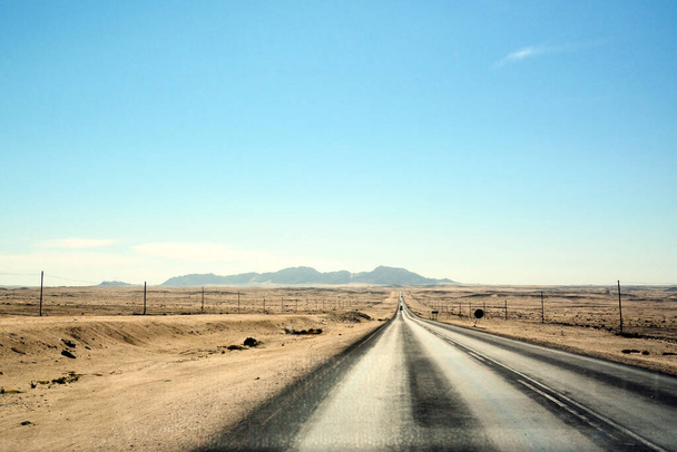 Ένας πολύ μακρύς δρόμος μπροστά στην έρημο. Άμμος και στύλοι ηλεκτροδότησης στα πλάγια. Μικρά βουνά είναι στο βάθος. Ζεστό κλίμα και έρημο τοπίο - Φωτογραφία, εικόνα