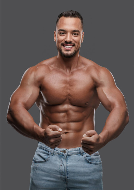 Incredibly attractive male model with a well-toned physique flashes a bright smile as he poses shirtless against a simple grey background, exuding charisma and magnetism - Fotó, kép