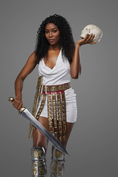 Prachtige donkere huid model heeft een witte Griekse tuniek met een sierlijke riem en kanen, met een gladius zwaard en schedel op een neutrale grijze achtergrond - Foto, afbeelding