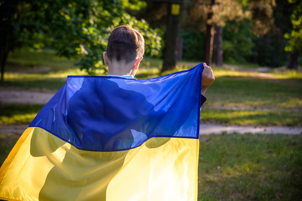 ウクライナのために祈る。夏の公園を走るウクライナの旗を持つ少年。国旗を振って平和を祈る小さな子供。独立記念日を祝う幸せな子供. - 写真・画像