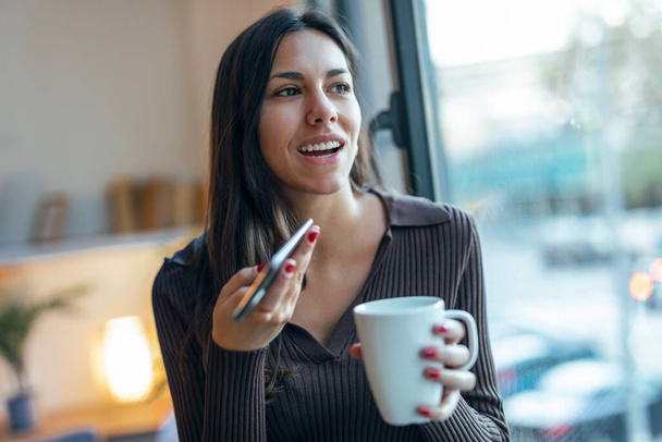 Shot of pretty young woman using her mobile phone while drinking coffee near to the window in the living room at home - 写真・画像