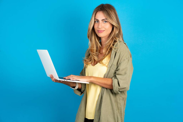 Adorable hermosa mujer rubia vistiendo una camiseta sobre fondo azul sosteniendo un dispositivo moderno - Foto, Imagen