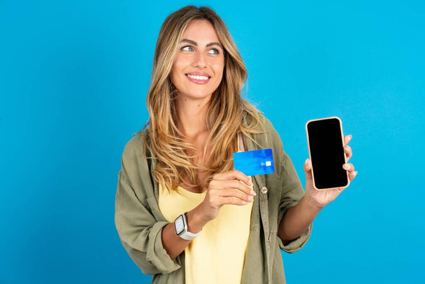 молода красива блондинка, одягнена в підшлункову залозу на синьому фоні, тримає банківську карту сучасний пристрій, виглядає порожнім простором - Фото, зображення