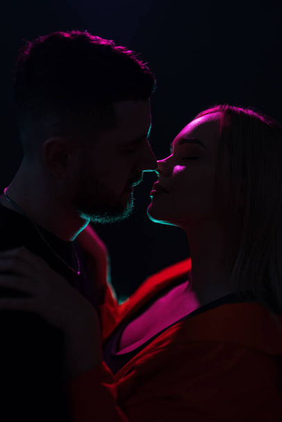 Πλευρική άποψη του νεαρού ζευγαριού φιλιούνται με κλειστά μάτια ενώ στέκονται στο σκοτάδι με φωτισμό νέον - Φωτογραφία, εικόνα