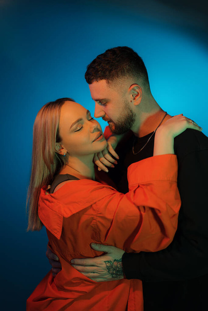 Vista laterale della giovane coppia innamorata vestita con abiti casual che si toccano mentre si trova in una stanza buia con illuminazione al neon blu - Foto, immagini