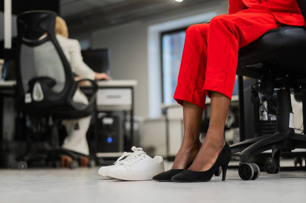 Mujer africana cambiando de tacones altos a zapatillas de deporte en la oficina. Piernas cansadas - Foto, Imagen
