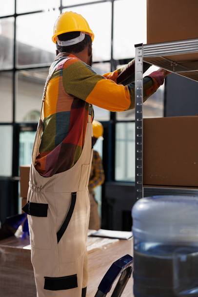 African american man standing on ladder and searching parcel on shelf in industrial warehouse. Storehouse worker wearing hard hat taking cardboard box for client order packing in stockroom - Foto, afbeelding
