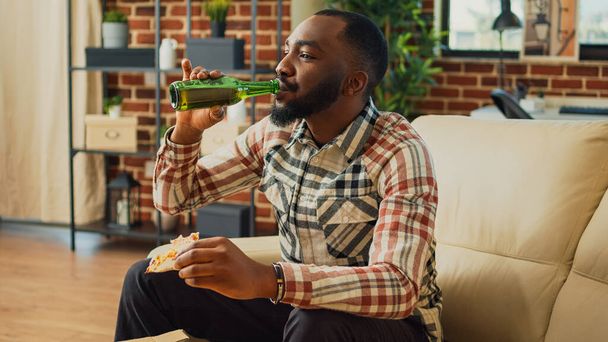 Young man eating slice of pizza at television on couch, enjoying takeout meal and bottle of beer at home. Modern person serving takeaway dinner and alcohol, watching film on tv. - Photo, Image