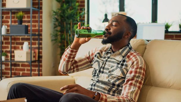 African american person eating burger and fries on sofa, having dinner with fast food and bottle of alcohol at home. Young man feeling happy watching action movie or show, having fun. - Foto, imagen
