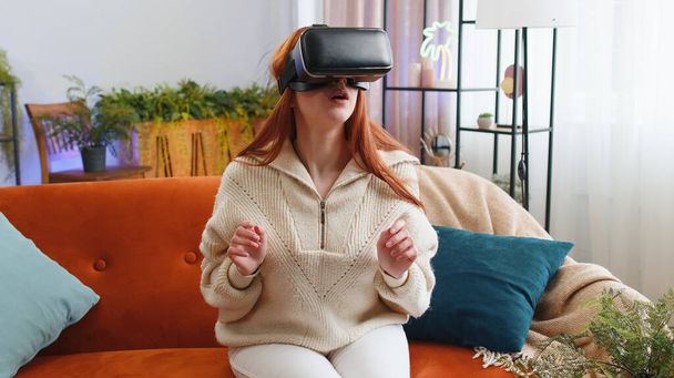 Redhead woman using virtual reality futuristic technology VR app headset helmet to play simulation 3D 360 video game, watching film movie at modern home apartment. Girl in goggles sitting on couch - Photo, Image