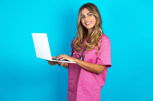 Очаровательная красивая женщина-врач стоит на синем фоне студии, держа в руках современное устройство - Фото, изображение
