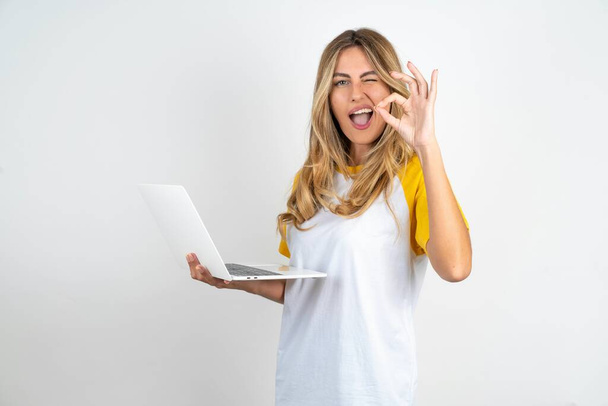 Приваблива весела кваліфікована красива жінка в футболній футболці на білому тлі з використанням ноутбука, що демонструє добре видно і підморгнув - Фото, зображення