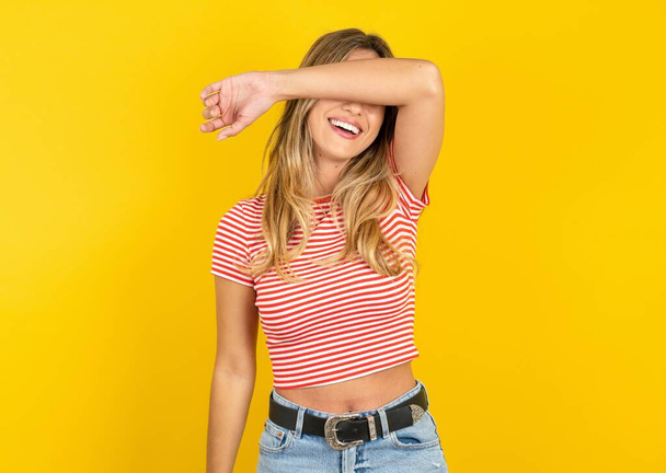 Piękna blondynka ubrana w koszulkę w paski na żółtym tle studia pokrywająca oczy uśmiechniętym ramieniem wesołym i zabawnym. Koncepcja niewidomych. - Zdjęcie, obraz