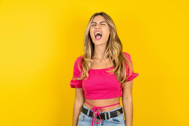 Angry and mad blonde young woman wearing pink crop top on yellow background screaming frustrated and furious, shouting with anger. Rage and aggressive concept.  - Photo, Image