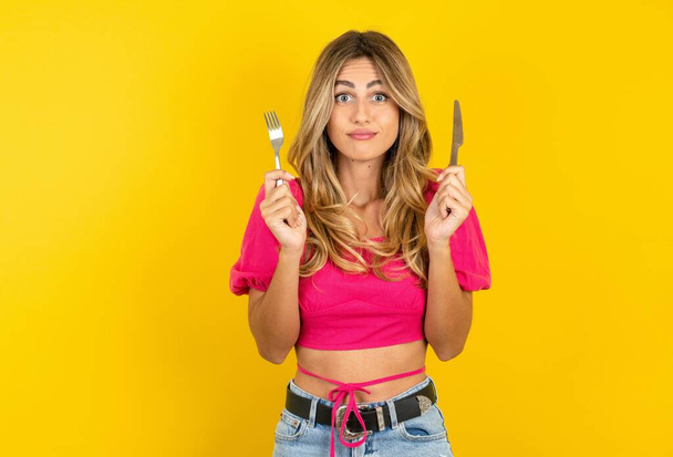 Hungry blonde young woman wearing pink crop top on yellow background holding in hand fork knife want tasty yummy pizza pie   - Photo, Image