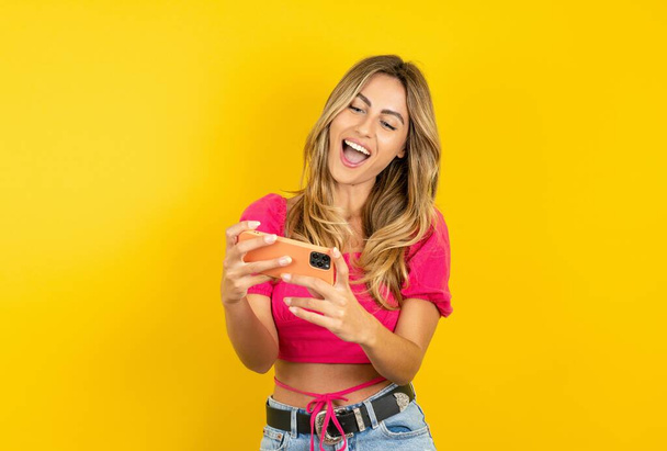 Portrait of an excited blonde young woman wearing pink crop top on yellow background playing games on mobile phone. - Foto, Bild