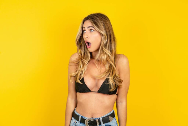 Young blonde woman on vacation wearing bikini over yellow background stares aside with wondered expression has speechless expression. Embarrassed model looks in surprise - Foto, Imagen