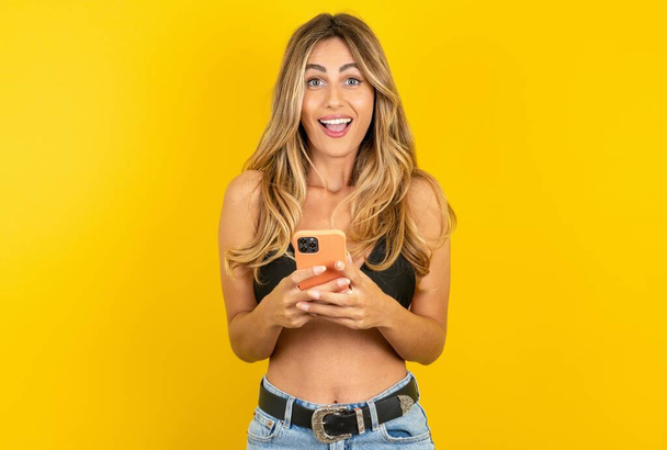 Schöne junge blonde Frau im Bikini vor gelbem Hintergrund hält Handy in den Händen und freut sich über positive Nachrichten, nutzt modernes Mobiltelefon - Foto, Bild