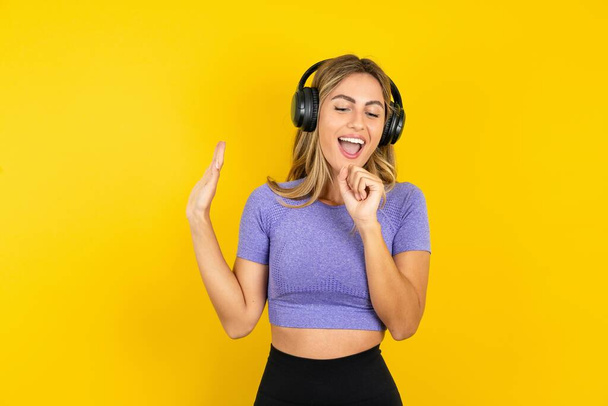 Happy young blonde woman wearing sportswear over yellow studio background sings favorite song keeps hand near mouth as if microphone wears wireless headphones, listens music  - Photo, image