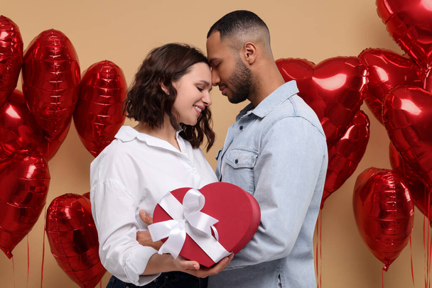 Lovely couple with gift box near heart shaped air balloons on beige background. Valentine's day celebration - Foto, afbeelding