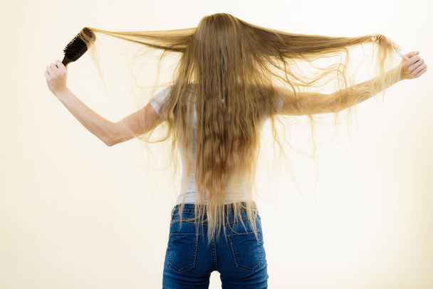 Blonde woman with brush combing her very long hair. Teenage girl with bad hair care. Back view - Фото, изображение