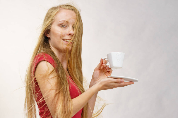 Blonde girl with blowing long hair holds heart shaped coffee cup, on grey - Photo, image