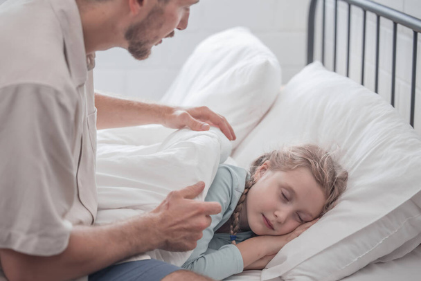 Father singing his daughter to sleep, providing comfort, support, and security. Focusing on fulfilling both physical and emotional needs. Kids sometimes act happy while pretending to fall asleep. - Zdjęcie, obraz