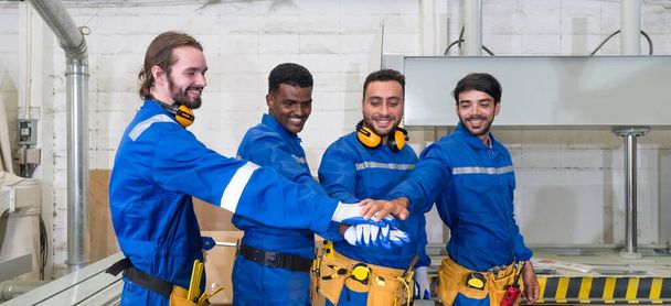 Group of technician in blue mechanic jumpsuit, ear muff and yellow tool belts join hand together in front of polywood cold press machine. Production line atmosphere in furniture factory - Photo, Image