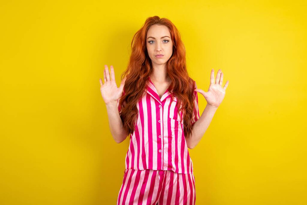 Ernsthafte rothaarige Frau im rosafarbenen Pyjama über gelbem Studiohintergrund zieht Handflächen in Richtung Kamera, macht Stoppgeste, bittet, die Emotionen zu kontrollieren und nicht nervös zu sein - Foto, Bild