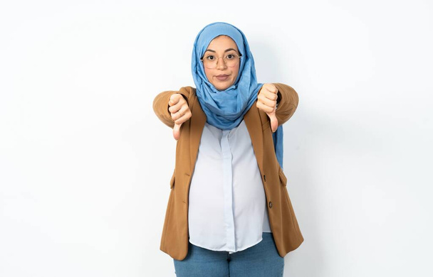 Mulher grávida muçulmana vestindo hijab sendo chateado mostrando polegar para baixo com duas mãos. Conceito de antipatia. - Foto, Imagem