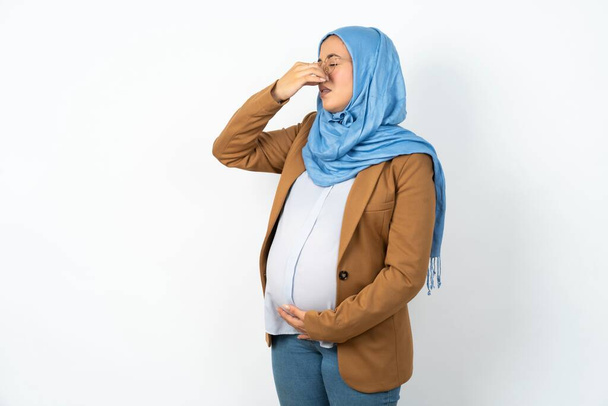moslim zwangere vrouw draagt hijab ruiken iets stinkend en walgelijk, ondraaglijke geur, houden adem met vingers op de neus. Slechte geur - Foto, afbeelding