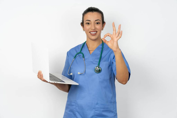 Pozitív arab orvos nő visel kék egyenruha hold vezeték nélküli netbook kéz ujjak mutatják okey szimbólum - Fotó, kép