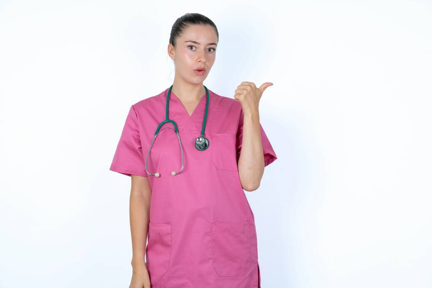 Choqué femme caucasienne médecin en uniforme rose avec des points de stéthoscope avec le pouce loin, indique quelque chose. Regarde-moi ça. Concept de publicité. - Photo, image