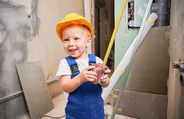 Szczęśliwy chłopiec budowlany pracownik trzyma taśmę pomiarową i uśmiecha się stojąc w mieszkaniu w trakcie remontu. Wesoły dzieciak z narzędziem pomiarowym w kasku ochronnym i kombinezonie roboczym. - Zdjęcie, obraz