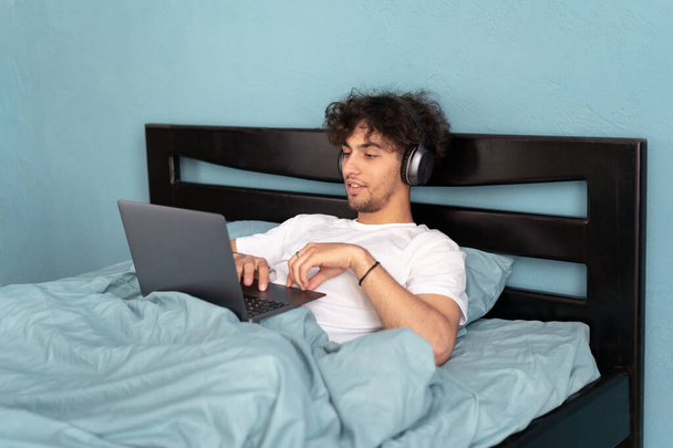 Handsome man working or studying remotely while lying in bed, freelancer working from home, web designer wearing headphones creating website in bedroom on weekend. Copy space - 写真・画像
