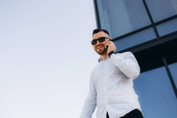 Un joven hombre de negocios guapo parado frente al edificio de oficinas con su portátil en la mano mientras habla por teléfono
 - Foto, imagen