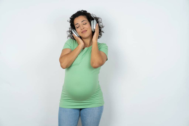 Pleased pregnant woman enjoys listening pleasant melody keeps hands on stereo headphones with her eyes closed. Spending free time with music - Fotografie, Obrázek