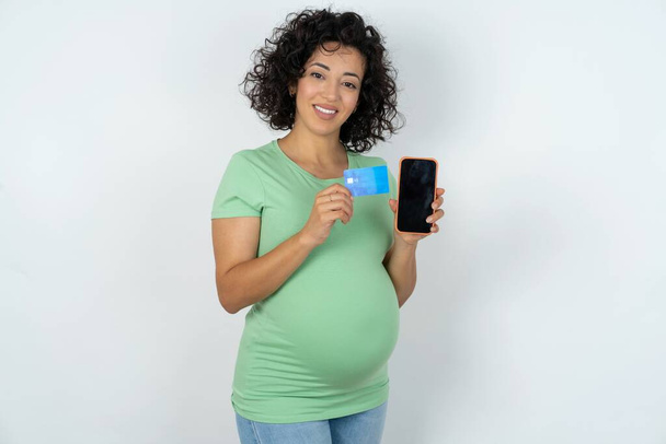 Foto van een schattige zwangere vrouw met een creditcard en smartphone. Gereserveerd voor online aankopen - Foto, afbeelding