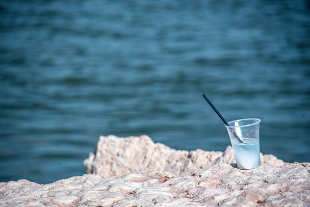Πλαστικό cocktail κύπελλο με ένα καλαμάκι εγκαταλειμμένο στα βράχια δίπλα στη θάλασσα. - Φωτογραφία, εικόνα