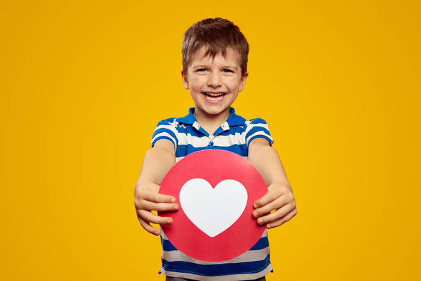 Adorable niño en polo de rayas azules, demostrando el icono del corazón para las redes sociales mientras está de pie sobre el fondo amarillo - Foto, Imagen