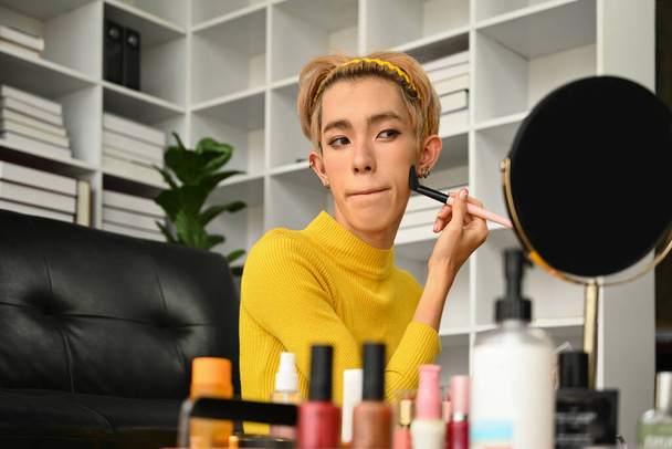 Genç eşcinsel adam oturma odasında günlük makyaj yapıyor, temel temeli süngerle yüzüne uyguluyor. LGBTQ, yaşam tarzı ve makyaj konsepti. - Fotoğraf, Görsel