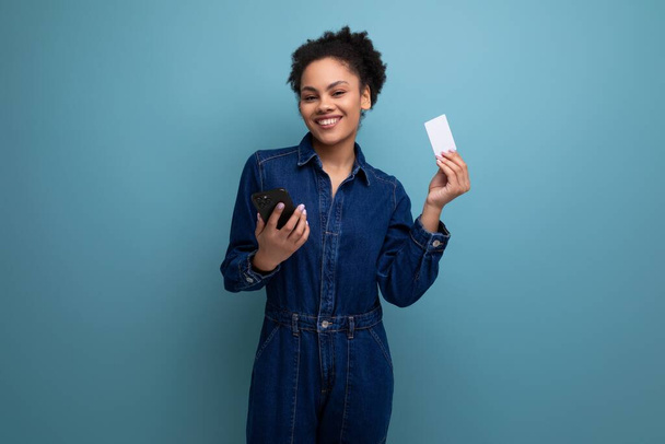 młody uroczy brunetka latynoski kobieta w niebieski dżinsowy garnitur gospodarstwa karty kredytowej makieta i smartphone. - Zdjęcie, obraz